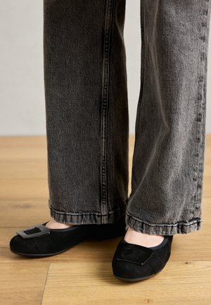 Schwarze flache Schuhe mit einer strukturierten Oberfläche, versehen mit einer quadratischen metallischen Schnalle. Getragen mit lässigen grauen Jeans auf einem Holzboden.