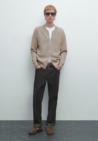 Cardigan beige lavorato a maglia con chiusura zip, t-shirt bianca, pantaloni scuri e mocassini marroni. Modella che indossa occhiali da sole scuri. Sfondo semplice.