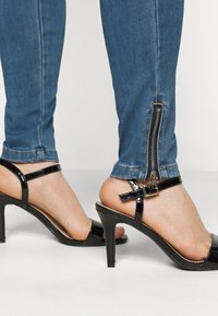 Sandalias negras de charol con tacón alto que presentan una correa alrededor del tobillo y una hebilla delicada. Vaqueros en un lavado azul claro con una cremallera lateral.