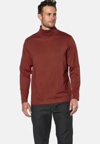 Langärmliger Rollkragenpulli aus rostfarbener Baumwolle mit glatter Textur und gesticktem Logo auf der Brust. Kombiniert mit dunkelgrauen Hosen.