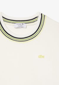 T-shirt crema con scollatura a coste caratterizzata da strisce nere e verdi. Logo ricamato giallo sul davanti. Vestibilità rilassata.