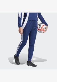 Marineblauer Trainingsanzug mit weißen Akzentstreifen und drei weißen Streifen an den Seiten, getragen mit schwarzen Fußballschuhen, während er einen Fußball hält.