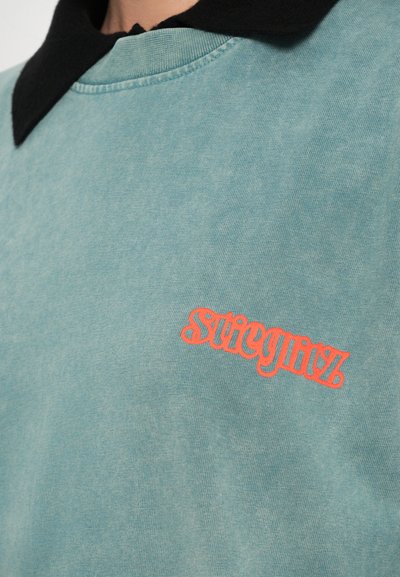 Tirkīza kokvilnas polo krekls ar melnu apkakli, kurā uz kreisā sāna ir izšūts spilgti oranžs teksts "streegitz". Mīksta tekstūra.