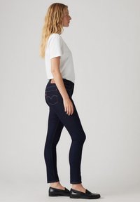 Jeans ajustados de mezclilla azul oscuro con costuras amarillas, combinados con una camiseta blanca y mocasines negros. Presenta un diseño en el bolsillo trasero.