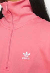 Růžová sportovní mikina na půl zip s vysokým límcem, vyrobená z měkké tkaniny, s vyšitým bílým logem Adidas na levé hrudi.