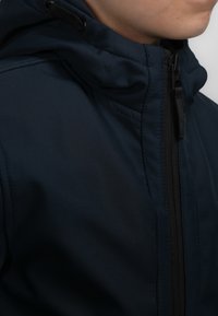 Námořnická modrá bunda s kapucí vyrobená z texturovaného materiálu, s plným zipem a černými detaily zipu, zobrazená ze strany, zvýrazňující kapuci a límec.