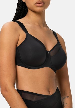 Nainen, joka käyttää mustaa alapaitaa ohuilla hihnoilla ja verkko-osiolla, nähdään hartioista vyötärölle yksinkertaista taustaa vasten.