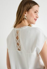 Blouse blanche à manches courtes, avec une finition texturée et un panneau arrière en dentelle à motifs floraux. Pas de fermetures ou d'embellissements visibles.