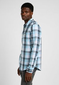 Koszula zapinana na guziki w jasnoniebieską i czarną kratę. Bawełnany materiał, długie rękawy i rozcięcia po bokach. Model ma na sobie szare dżinsy.