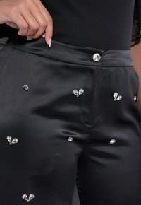 Černé saténové kalhoty s dekorativními kamínky ve tvaru srdce, hladkou texturou a zapínáním na knoflík v pase.