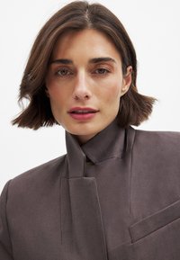 Brown tailored blazer with a high collar, structured design, and front pocket detail, made from smooth fabric with a matte finish.