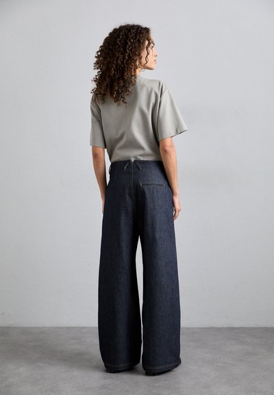 Chemise grise à manches courtes associée à un pantalon en denim foncé à jambe large, doté de poches latérales bien visibles et d'une poche arrière avec des accents de couture jaune.