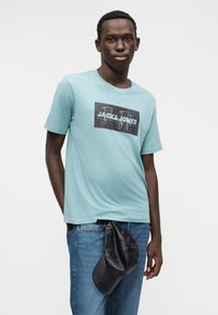 Ανοιχτό μπλε βαμβακερό t-shirt με κοντά μανίκια και μαύρο γραφικό λογότυπο. Συνδυασμένο με μπλε τζιν και μαύρη τσάντα καπέλο.