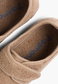 Zapatillas de tela en color beige con una suave textura de toalla. Cuentan con una correa de velcro y un logo azul visible en la plantilla. Diseño de punta redonda.