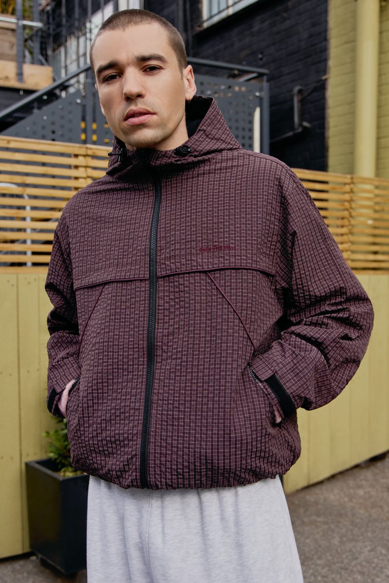 Giacca zip-up a scacchi bordeaux con cappuccio, dotata di tasche laterali e dettagli neri. Indossata con pantaloni grigi chiari, su uno sfondo di legno.