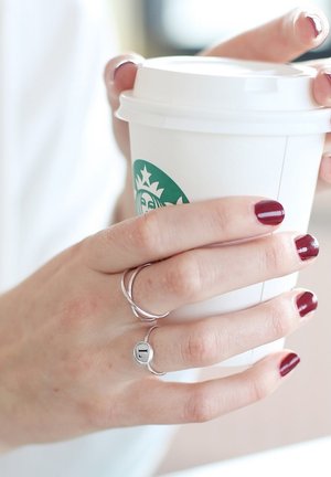 Weißer Pappbecher mit grünem Logo, der in einer Hand gehalten wird. Die Hand ist mit zwei ineinander verschlungenen silbernen Ringen und einem Ring mit dem Buchstaben "L" geschmückt.