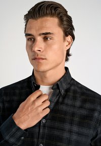 Jonge man met kort donker haar die een zwart geruit overhemd over een wit t-shirt draagt, met zijn rechterhand de kraag aanpast en naar links kijkt.