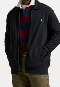 Man i svart zip-up jacka över en vinröd och marinblå randig skjorta, med olivgröna cargobyxor och ett bälte, händerna i jackfickorna.