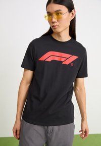 Black T-shirt with red stylized logo on the front, short sleeves, and crew neck. Worn with yellow sunglasses and gray pants, on a green surface.