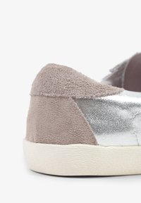 Zapatilla de ante gris y metalizada plateada con una suela texturizada en blanco roto y un panel plateado liso en contraste. Diseño de parte trasera redondeada.