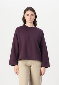 Felpa oversized viola con maniche a campana lunghe, scollatura rotonda, texture morbida e vestibilità rilassata; abbinata a pantaloni beige.