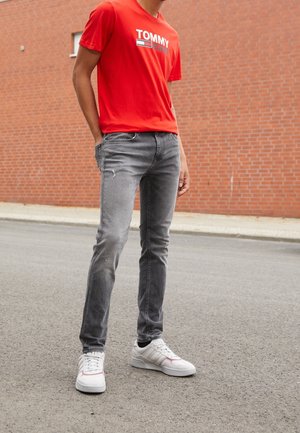 T-shirt en coton rouge avec le logo "TOMMY", associé à un jean en denim gris avec des détails usés, et des baskets blanches avec des accents rouges.