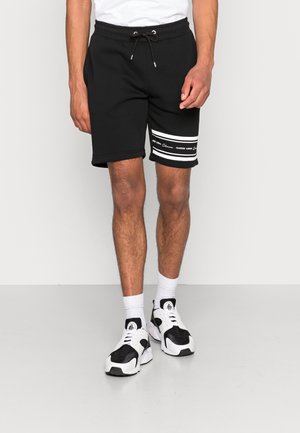 Mann trägt schwarze Shorts mit Kordelzug, weißen Streifen und Text am linken Bein, weiße Socken und schwarz-weiße Sneakers.