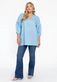 Lichtblauw, oversized blouse met een V-hals en pofmouwen; gecombineerd met uitlopende blauwe jeans en lichtblauwe sandalen met bandjes.