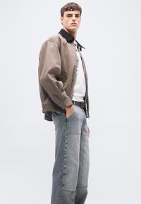 Νέος άνδρας στέκεται με τα χέρια στις τσέπες, φορώντας ένα καφέ μπουφάν με μαύρο γιακά, λευκό πουκάμισο και φαρδιά γκρι τζιν με σχέδια.