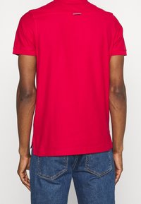 Polo rouge à manches courtes, tissu affichant une texture lisse, le dos présente une petite fente, associé à un jean en denim bleu.