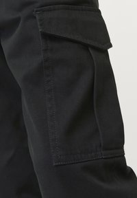 Pantalones cargo negros con un bolsillo lateral con solapa, costuras visibles y una textura de tela suave y duradera. Enfocados en la utilidad y los detalles de diseño.