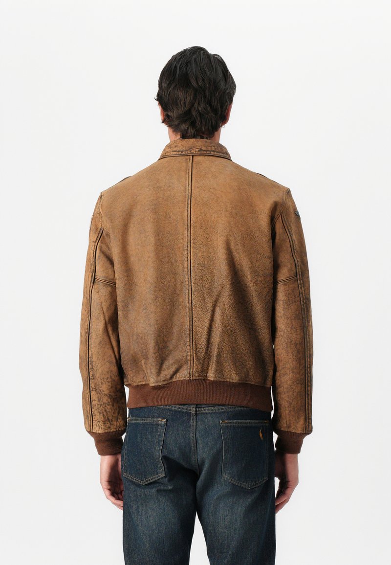 Giacca bomber in pelle marrone con finitura testurizzata, polsini e orlo a costine, e colletto classico, indossata sopra jeans blu scuro.