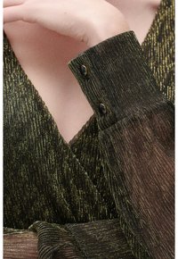 Tessuto di un abito lucido nero e oro con scollatura a V, maniche lunghe in trasparente e polsini con bottoni. Presenta un design avvolgente e un motivo testurizzato.