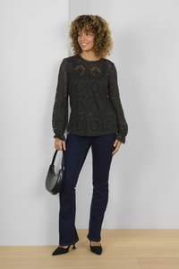 Top in pizzo nero con maniche lunghe e motivo floreale, abbinato a jeans svasati di un blu scuro. La modella tiene una borsa nera a forma curva.