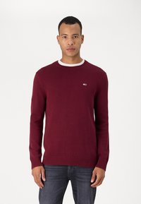 Pull en maille bordeaux avec un col rond, des poignets et un ourlet côtelés. Il comporte un petit logo sur la poitrine gauche. Porté par-dessus une chemise blanche.