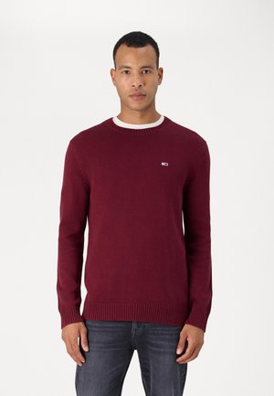 Suéter de punto burdeos con cuello redondo, puños y dobladillo acanalados. Presenta un pequeño logotipo en el lado izquierdo del pecho. Lleva puesto un camisa blanca debajo.