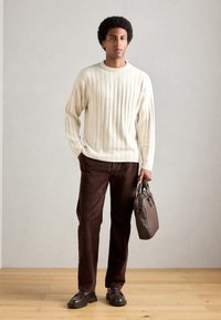 Lyscreme ribbet sweater, brune fløjlsbukser og sorte loafers. Holder en mørkebrun lædertaske. Enkel og struktureret design.