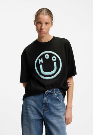 Kvinde med lyst brunt hår iført en sort oversized T-shirt med blåt "HUGO" smileylogo og løse blå jeans, stående mod en hvid baggrund.