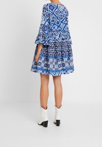 Modré vzorované šaty s volánkovými rukávy, které mají přiléhavý korzet a rozšířenou sukni, vyrobené z lehké tkaniny, vynikající s bílými kotníkovými botami.