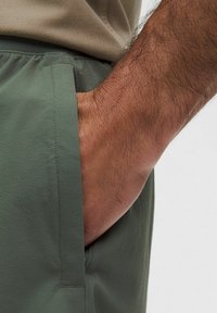 Täiskasvanu käsi toetub oliivirohelistes tekstuuriga lühikestes pükstes esimesse taskusse, beež särk on osaliselt nähtav vööst üle.