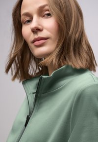 Giacca verde con cerniera frontale e colletto alto, dalla texture morbida, con dettaglio cerniera nero e design minimalista, indossata da una persona.