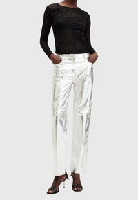 Personne portant un haut à manches longues en dentelle noire, un pantalon droit argenté brillant avec les mains dans les poches, et des sandales à talons hauts noires.