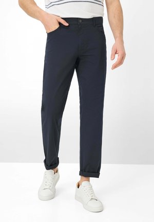 Mand iført marineblå bukser med optrukne ben, hvide sneakers og en hvid skjorte med blå striber, stående med den ene hånd i lommen.