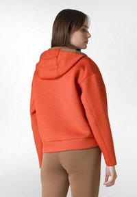 Orange sweatshirt med huva, med lös passform, nedsänkta axlar och bruna detaljer runt halsringningen. Slät textur, avslappnad design.