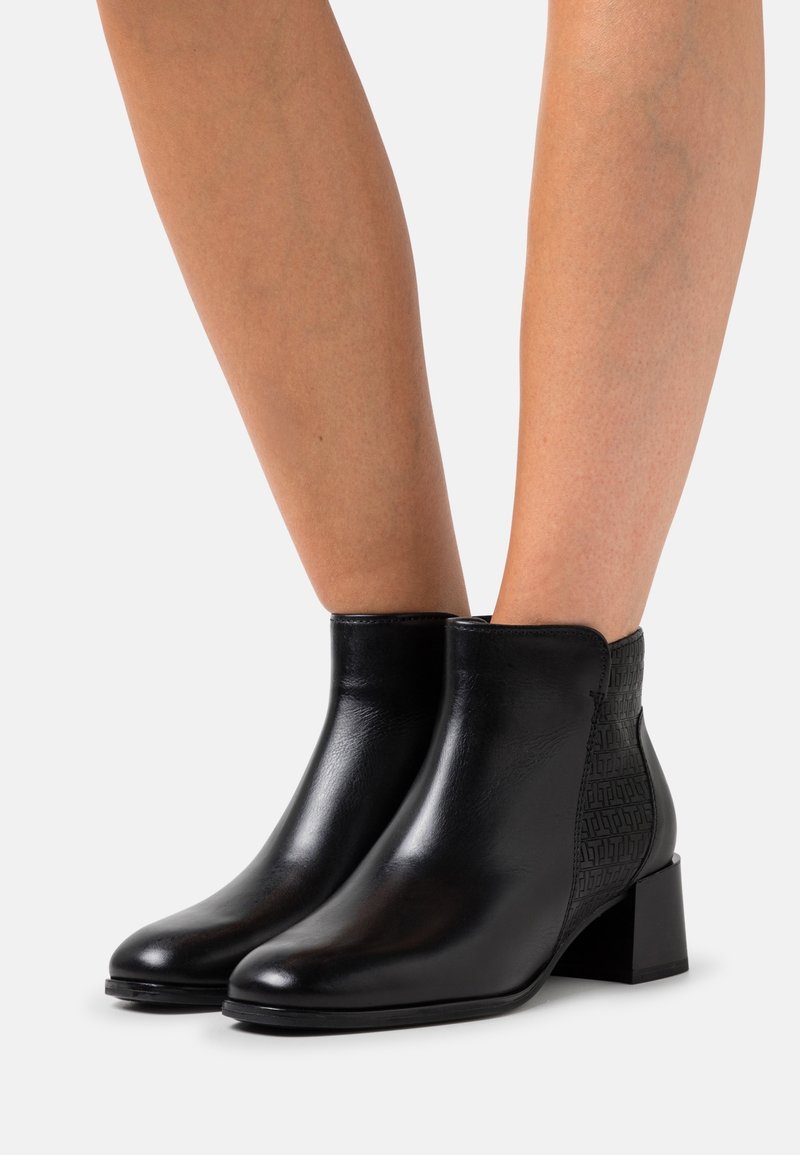 Las botas de tobillo negras presentan cuero suave y un panel texturizado con un patrón de logo. El tacón cuadrado mide aproximadamente 3 cm de altura.