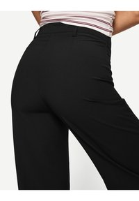 Svarta vida byxor med en slät textur, med midja i normal höjd och bakficka, visade från en sidovinkel.