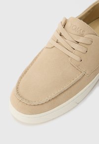 Beige velúr sneaker fűzővel, látható varrásokkal és fehér gumitalppal. Az oldalán lévő márkajelzés dombornyomott szöveget tartalmaz.