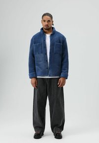 Blauwe fleecejas met een gestructureerd oppervlak, voorzien van twee voorzakken, knoopsluiting en een ontspannen pasvorm, gecombineerd met grijze jeans met wijde pijpen.