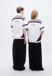 Λευκά σακάκια με κοντά μανίκια που διαθέτουν γεωμετρικά σχέδια σε τιρκουάζ και μαρόν, με μαύρες λεπτομέρειες και ελαστική μέση, φορεμένα με άνετο μαύρο παντελόνι.