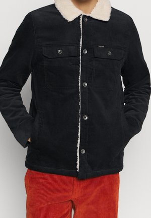 Schwarze Cordjacke mit weißem Lammfellkragen und silbernen Knöpfen, getragen über roten Cordhosen, Hände in den Taschen.